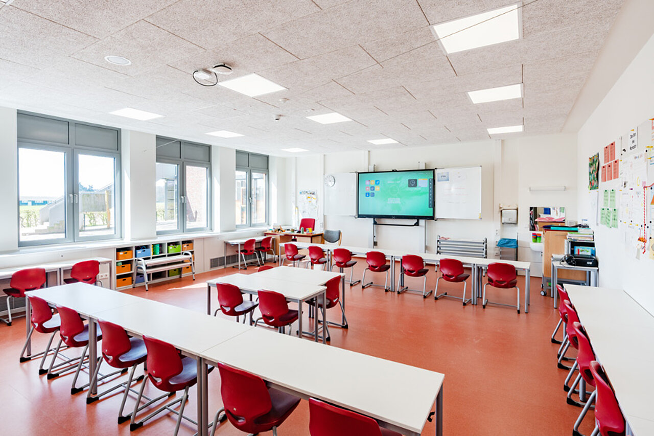 Klassenraum mit Tischen, roten Stühlen, Akustikdecke und digitaler Tafel